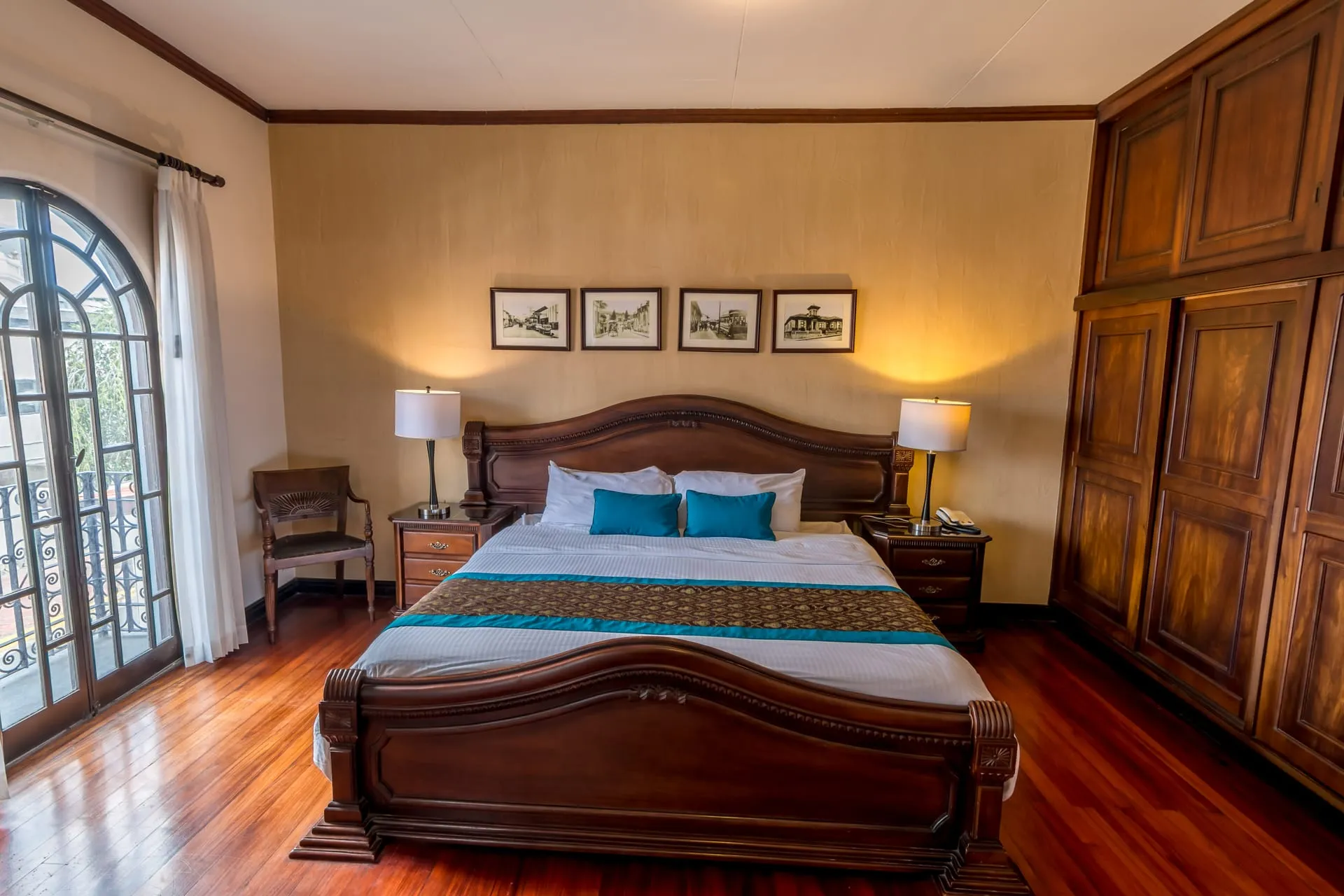 Suite Junior con cama doble extragrande de cabecera tallada en madera, piso de madera oscura y puerta-balcón de hierro forjado
